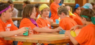 Group of children in orange tshirts and coloured buffs sitting at a camp table
