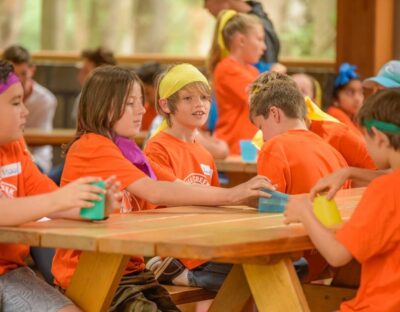 Group of children in orange tshirts and coloured buffs sitting at a camp table