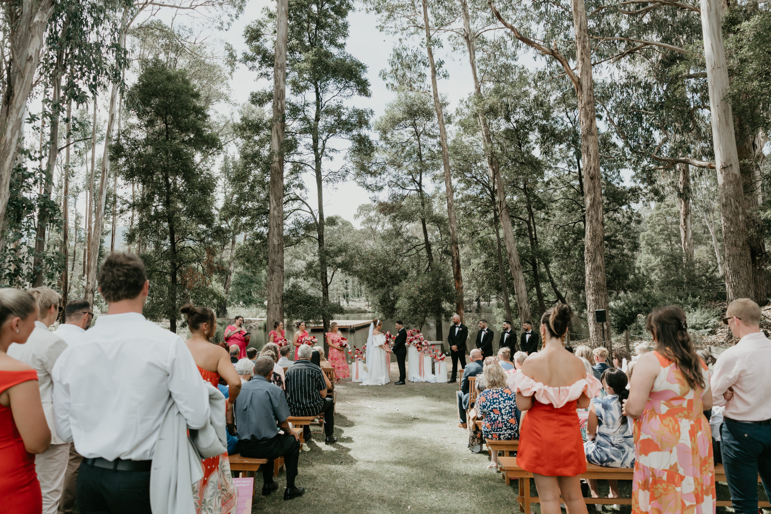 Guests gathering in pews to witness a wedding ceremony outdoors beneath the trees